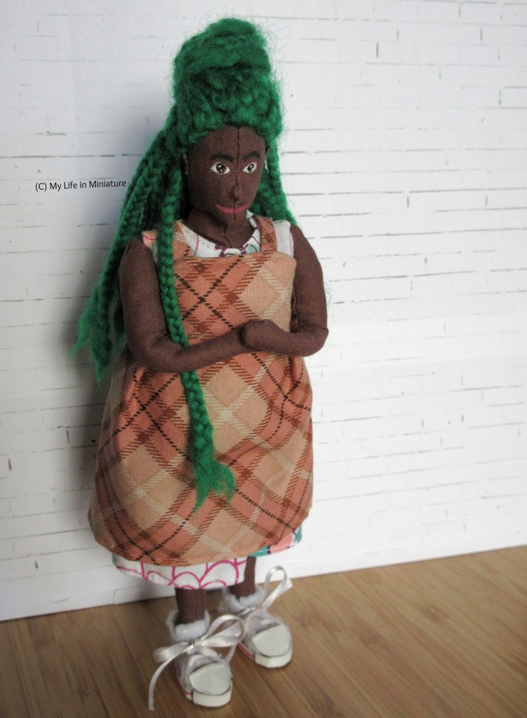 Hazel stands against a white brick background, wearing the peach pinafore over her white dress. She has one leg extended in front of her, and is looking down at the dress. Her hands are clasped at her waist. One braid falls over her shoulder. 