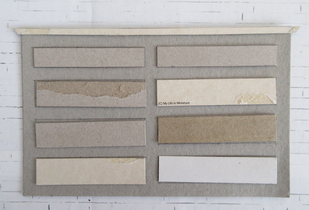 The flat cardboard front of a chest of drawers lays on a white brick background. There are eight drawers on the front and a decorative strip of cardboard along the top. All the cardboard is in its plain grey/brown unfinished state, and none of it is glued down. 