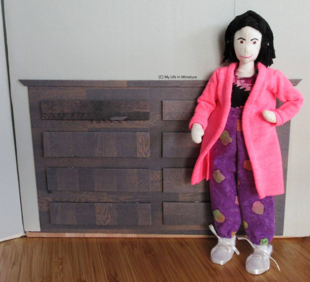 Tiffany stands against a wall in Needle & Thread, one hand on her hip. The other elbow is resting on an open drawer of the dark wood chest of drawers behind her. The drawers come to just below her shoulders, and there are eight drawers in the unit.