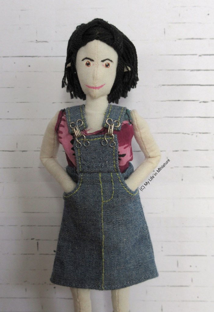 Tiffany stands against a white brick background, smiling at the camera with both hands in her pinafore pockets. The front panel of the pinafore is smaller, and the straps are now a good length.