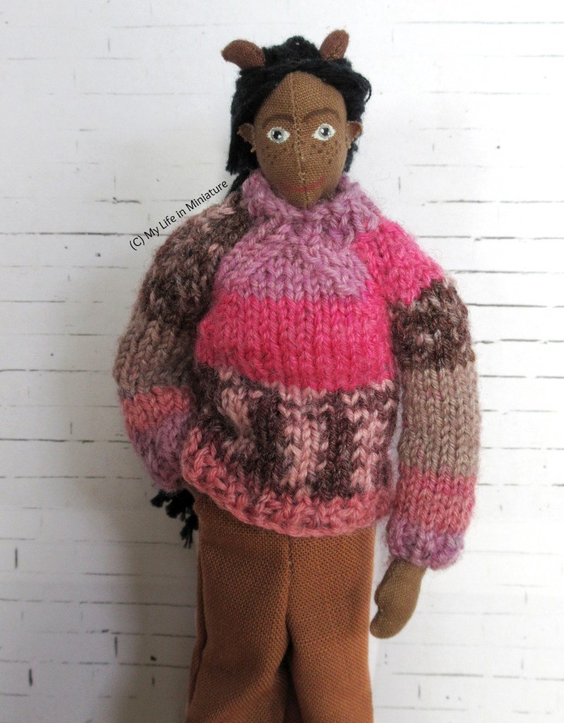 Petra stands against a white brick background, smiling at the camera. She wears the brown pants with a pink knitted jumper, one hand in her pant pocket.