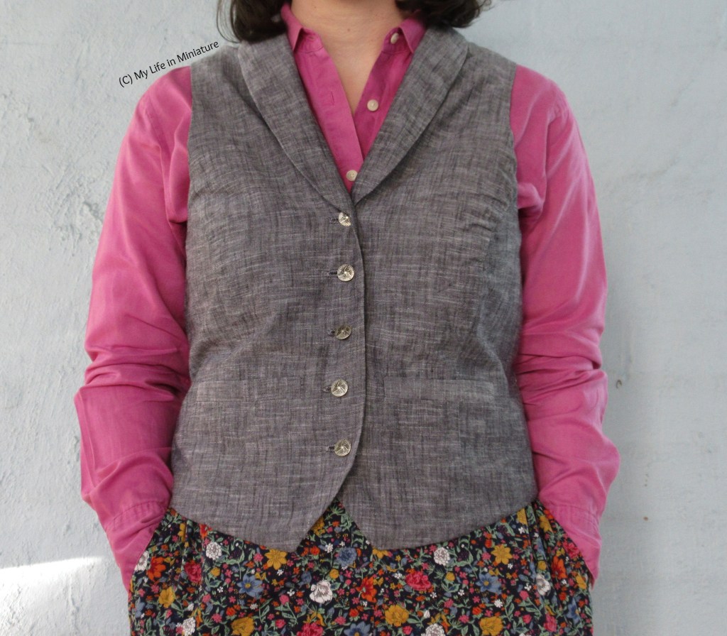 The author stands against a plain grey wall, photographed from shoulders down. She wears a plain grey waistcoat with a shawl collar over a pink linen shirt and floral pants. Her hands are in her pockets. 
