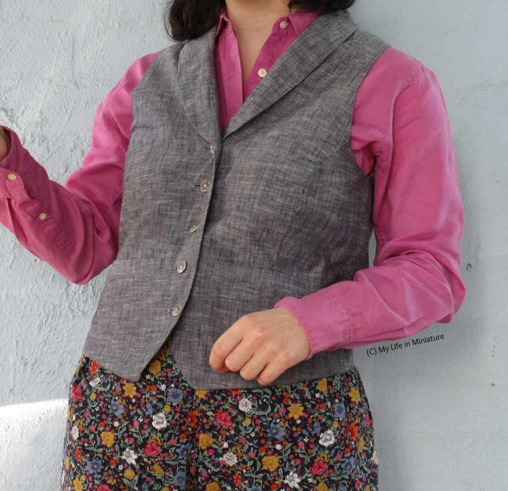The author stands against a plain white wall, photographed from the shoulders down. She wears the grey waistcoat, pink shirt, and floral pants. One arm is raised, hand cropped out of the image, while the other is held at her waist. Her body is held at an angle.