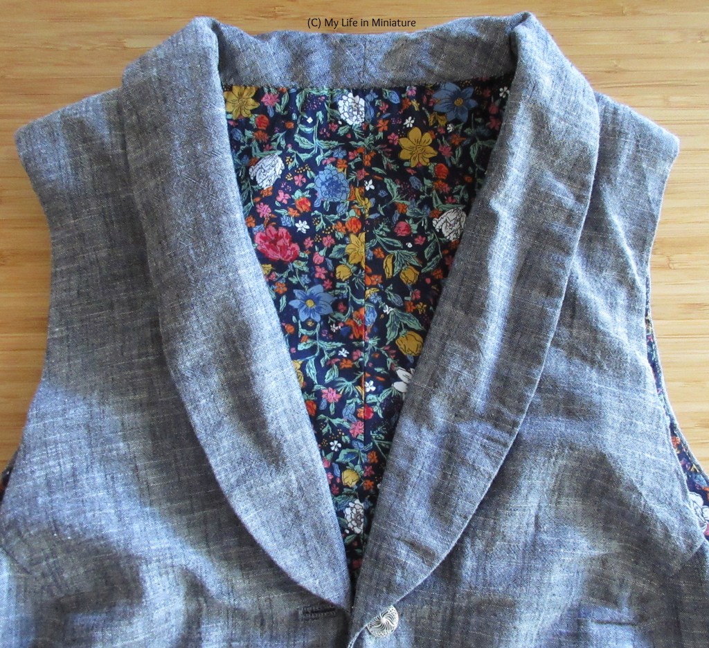 The plain grey waistcoat is laid flat on a wood surface. The image is cropped to the top half of the waistcoat, where the floral lining at the back can be seen through the shawl collar neckline. 