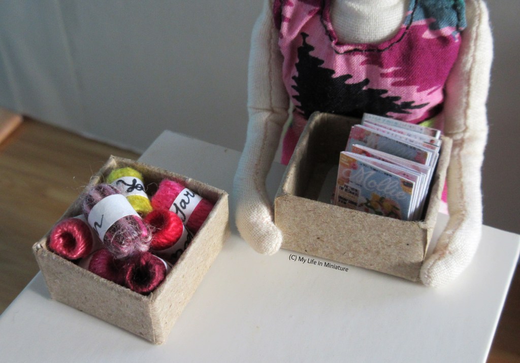 Tiffany places one of the brown baskets onto Needle & Thread's workbench, accompanying the one already there. The basket in her arms has craft magazines in, and the other basket is full of yarn skeins.