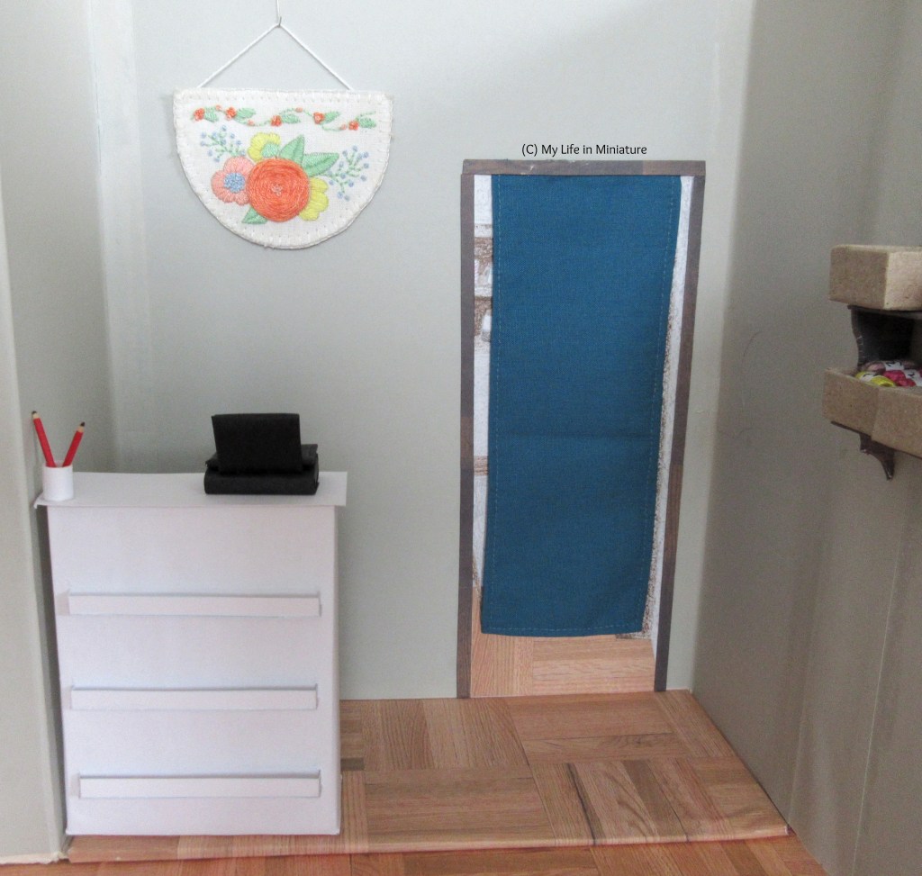 Interior of Needle & Thread. In the foreground is the white front counter, with cash register and two pencils on it. On the back wall is a teal-curtained doorway and a semi-circular embroidery. The curtain covers most of the doorway.