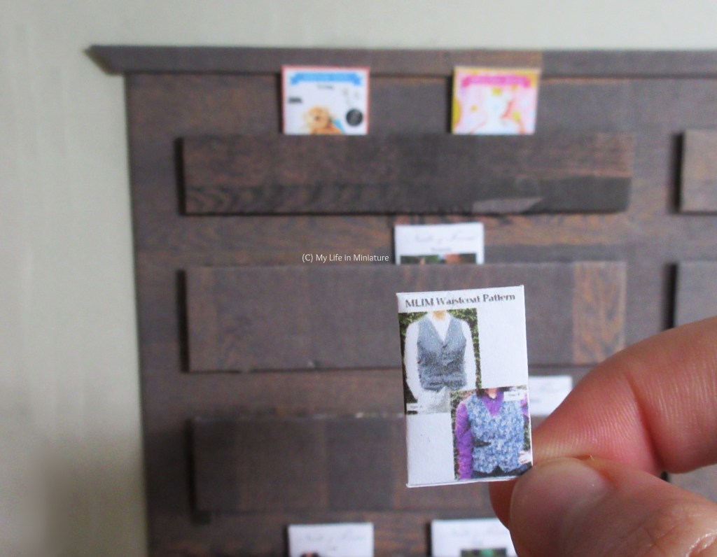 The author's hand holds a miniature version of the pattern pouch in front of the pattern drawers in Needle & Thread. 