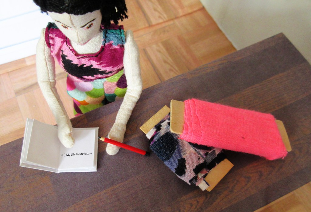 Overhead shot of the large table in Needle & Thread. The surface is now dark brown wood, and on it are two bolts of fabric and an open sewing book that Tiffany's hand holds open. Her other hand holds a pencil that is tapping the table. 