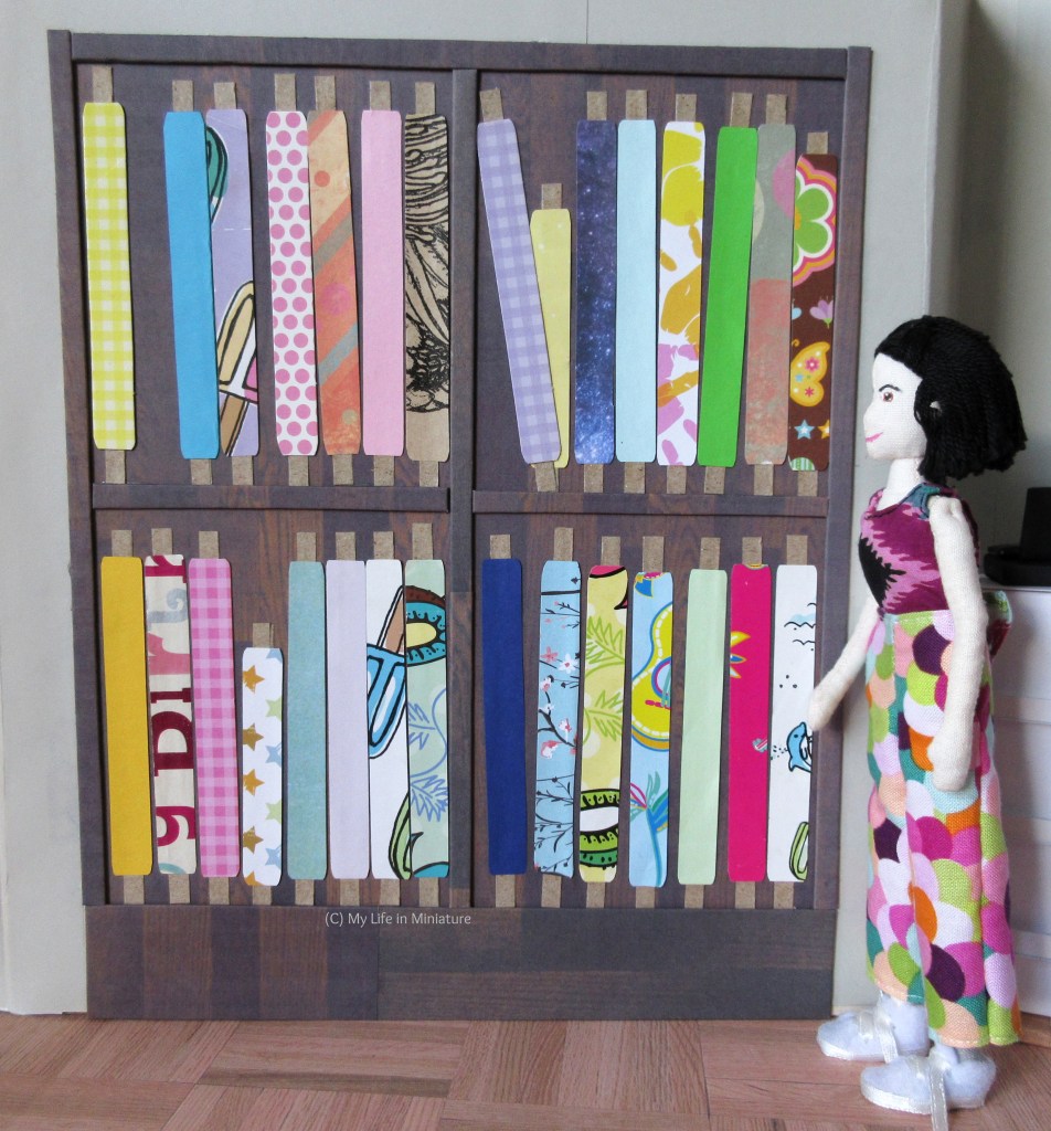 Tiffany stands beside the fabric display in Needle & Thread, so all the fabric can be seen. She wears a black, purple, and teal top with colourful pants, and looks at the display.
