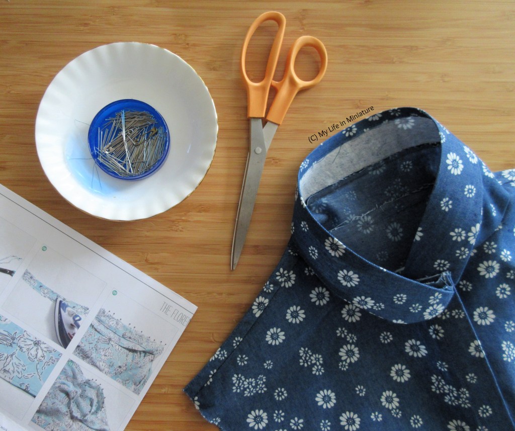 Flatlay shot of a bodice, some sewing instructions, a dish with a pin tin on, and a pair of fabric shears. The collar and shoulders of the bodice is visible, in a blue fabric with white daisies on. All items are on a wood background.
