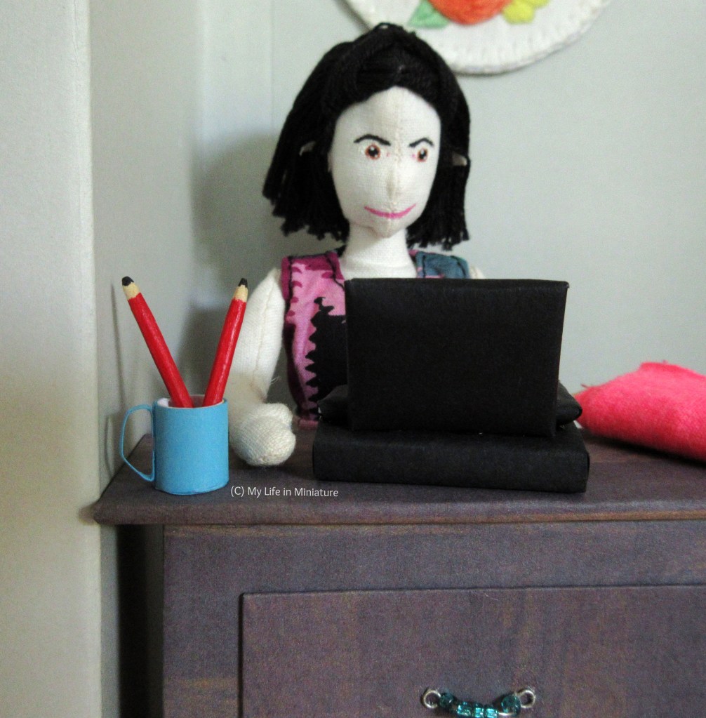 Tiffany stands at the front counter, using the cash register. The focus, however, is on the bright blue mug with two pencils in. Beside the cash register on the counter is a bolt of fabric.