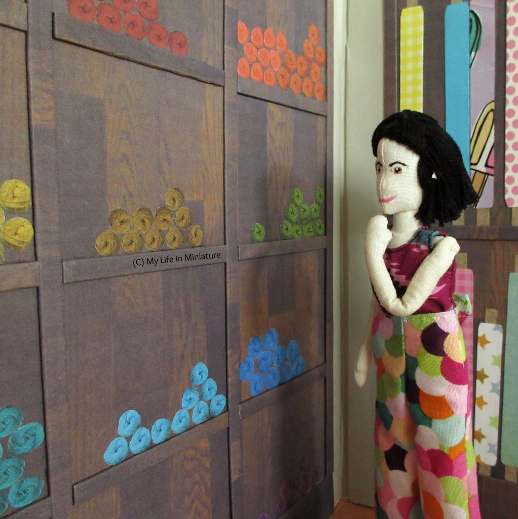 Tiffnay stands beside the yarn wall in Needle & Thread, looking at it with a thoughtful hand to her chin. 