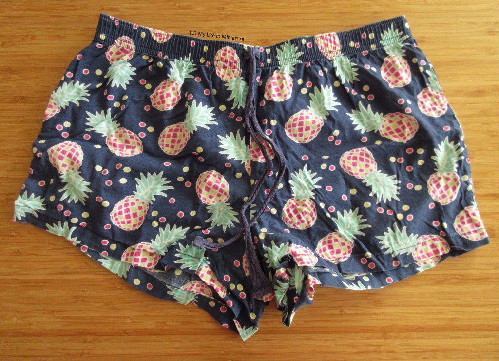 A pair of pyjama shorts is laid out flat on a wood surface. They are navy with green, yellow, and pink pineapples on them. There is a navy tie at the centre front of the elastic waistband.