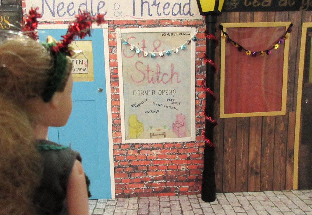 Over-the-shoulder shot of Sarah looking at the other side of the street, where Needle & Thread and Tea at Yaz's are decorated with garlands over their windows. The lamppost on this side has red tinsel wrapped around it.