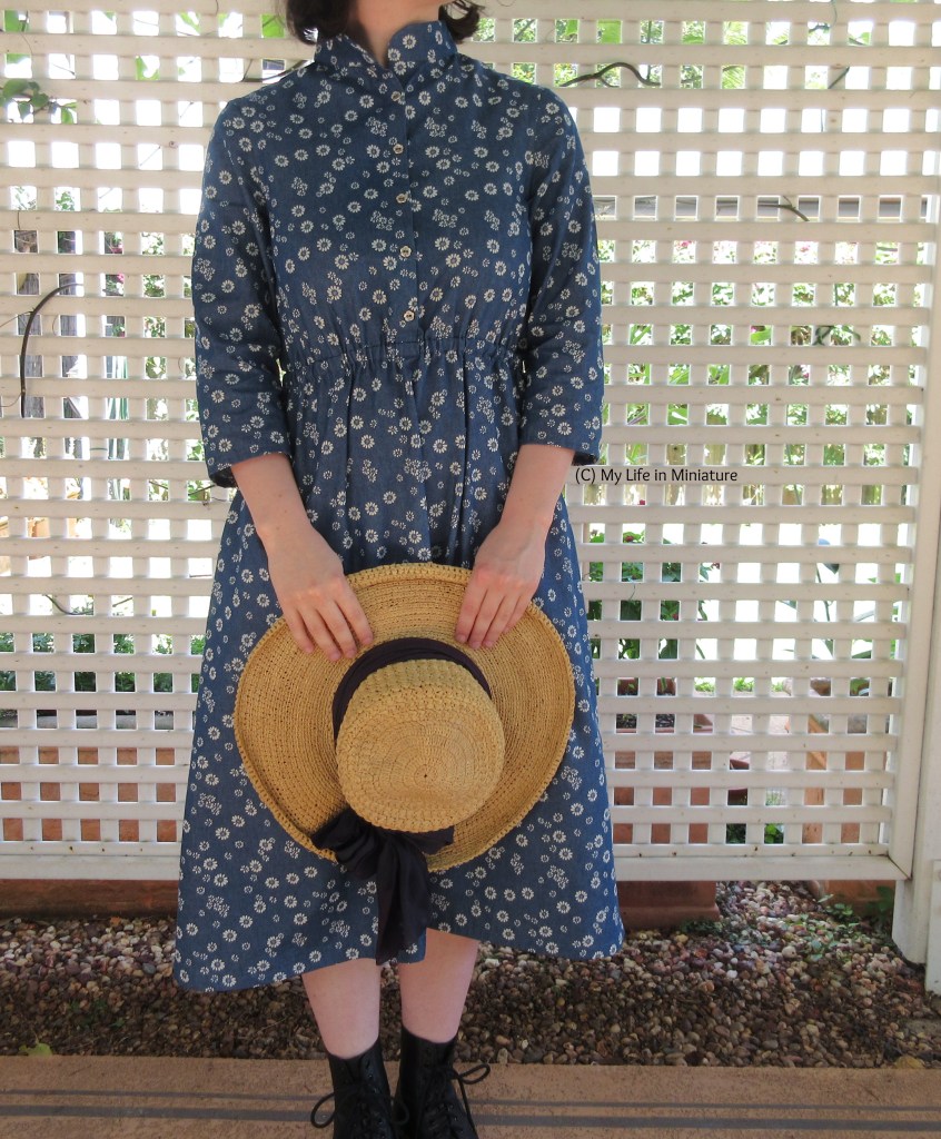 Almost full-body shot of the author against the lattice backdrop. She wears the blue daisy dress and black boots, and holds a straw hat in front of her. The hat has a thick navy blue band around it, tied in a bow. 
