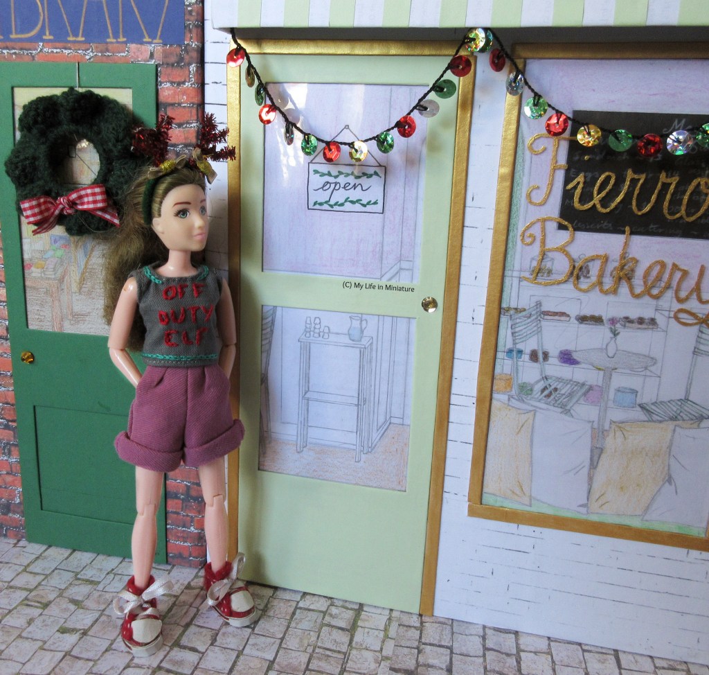 Sarah walks along the street, hands in pockets. She looks at Fierro's as she passes, which has a new sparkly garland hanging from its awning. The garland has a repeating pattern of red, green, then silver or gold sequins, crocheted together with black thread. 