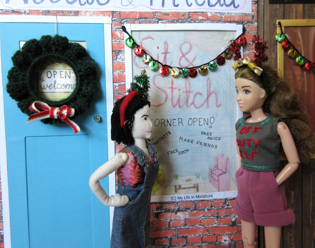 Sarah and Tiffany discuss the new decorations in the street. Tiffany has her hands on her hips, and wears a Christmas tree headband. Behind her, the wreath on Needle & Thread's door has a red, white, and gold bow on the centre bottom. 