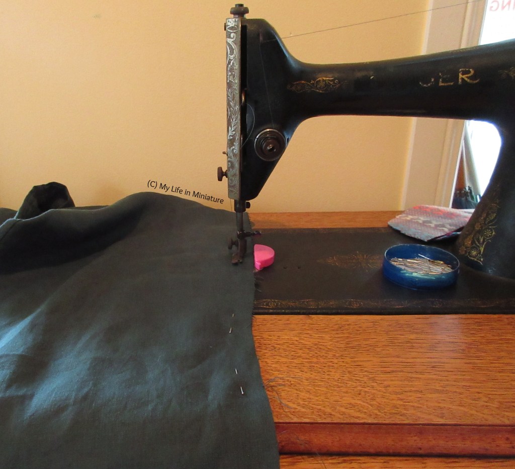 A large amount of dark greeen fabric is under the needle of an antique sewing machine. Nearby is a pin tin and a small scrap of thick fabric. The wall behind is pale yellow.