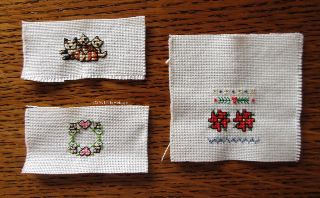 Three small unframed cross stitchings are laid flat on a wood background. Top left is a cat with three kittens, bottom left is a leafy wreath with flowers and hearts; right is a wintery sampler.