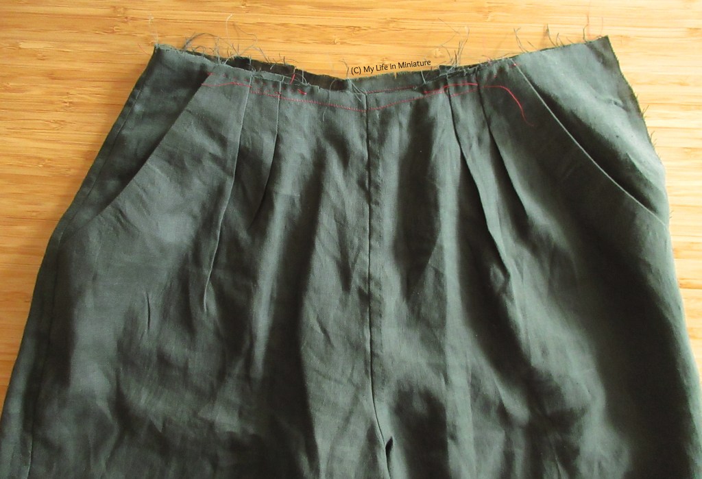 The top portion of a pair of unfinished dark green pants is laid flat on a wood background. The pants have pleats at the front, two diagonally-opening pockets, and the top edge is frayed and without a waistband.