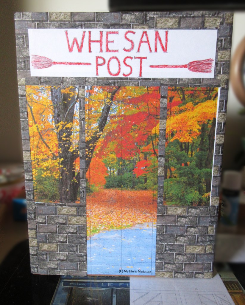 A cereal box stands on a work surface, facing the camera. It is completely covered in cardboard, and the cardobard is partially covered with dark brick paper. Across the top is a white sign with red writing that reads 'Whesan Post'. Also on the sign are two red broomsticks.