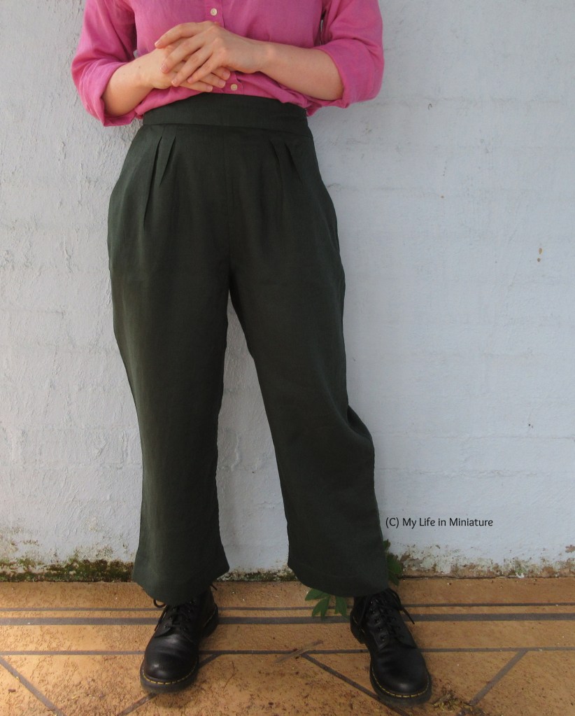 The author stands in front of a pale blue brick wall, wearing a pink linen shirt with dark green pants and black boots. Her hands are held in front of her body, just above the unelasticated waistband of the pants. 