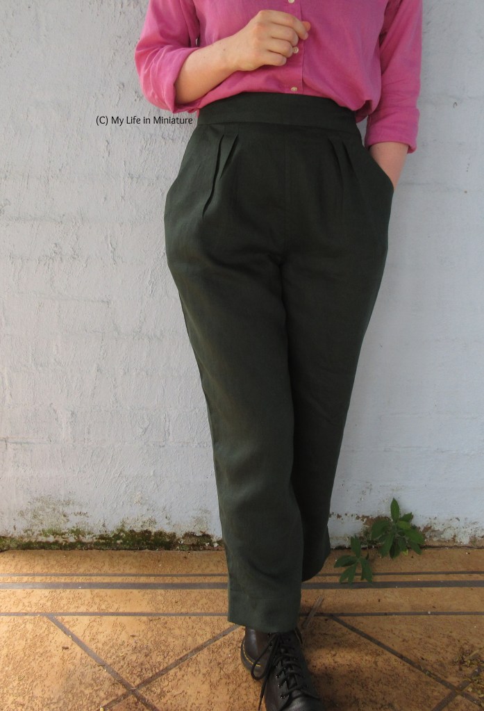 The author stands in front of a pale blue brick wall, wearing a pink linen shirt with dark green pants and black boots. One hand is in her pocket, the other is held in front of her body just above the waistband. Her feet are in line with each other, one heel resting on the toe of the other foot.