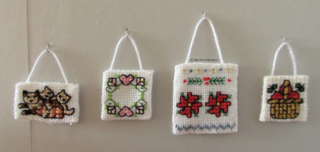 Close-up of the cross stitchings. Left to right they depict a cat with three kittens, a wreath with hearts and flowers, a wintery sampler, and a basket of apples. 