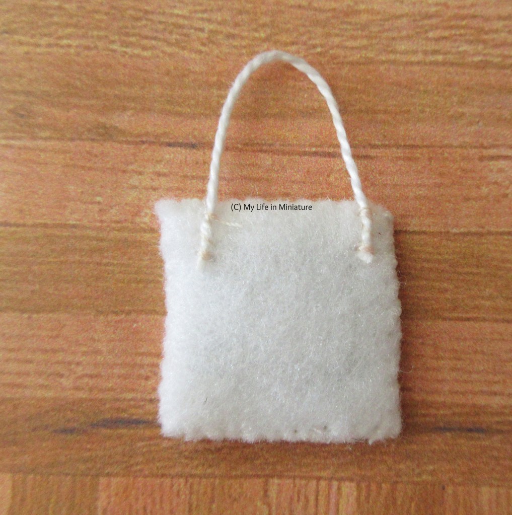 The reverse of one of the stitchings is on a wood background. It's a square of white felt with a string sewn to it in a loop at the top edge. 