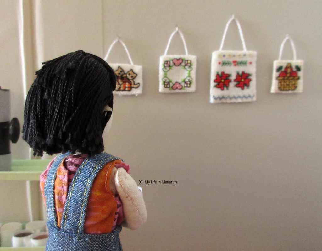 Tiffany stands with her back to the camera, looking at the stitchings. She wears a colourful shirt with a denim pinafore. 