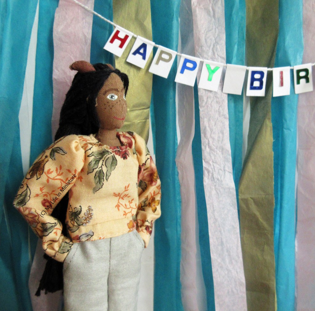 Petra stands in front of the party backdrop, hands in pockets. She wears a beige-yellow floral top with grey pants. She is looking at the 'Happy Birthday' banner, which is partially in 