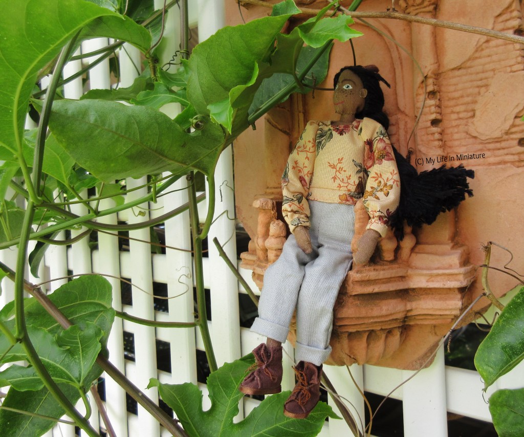 Petra sits in a terracotta garden ornament, designed to look like a crumbling classical Italian or Greek balcony. She has one hand in her lap, the other resting on the rail, and is looking up at the vines and leaves surrounding her. 