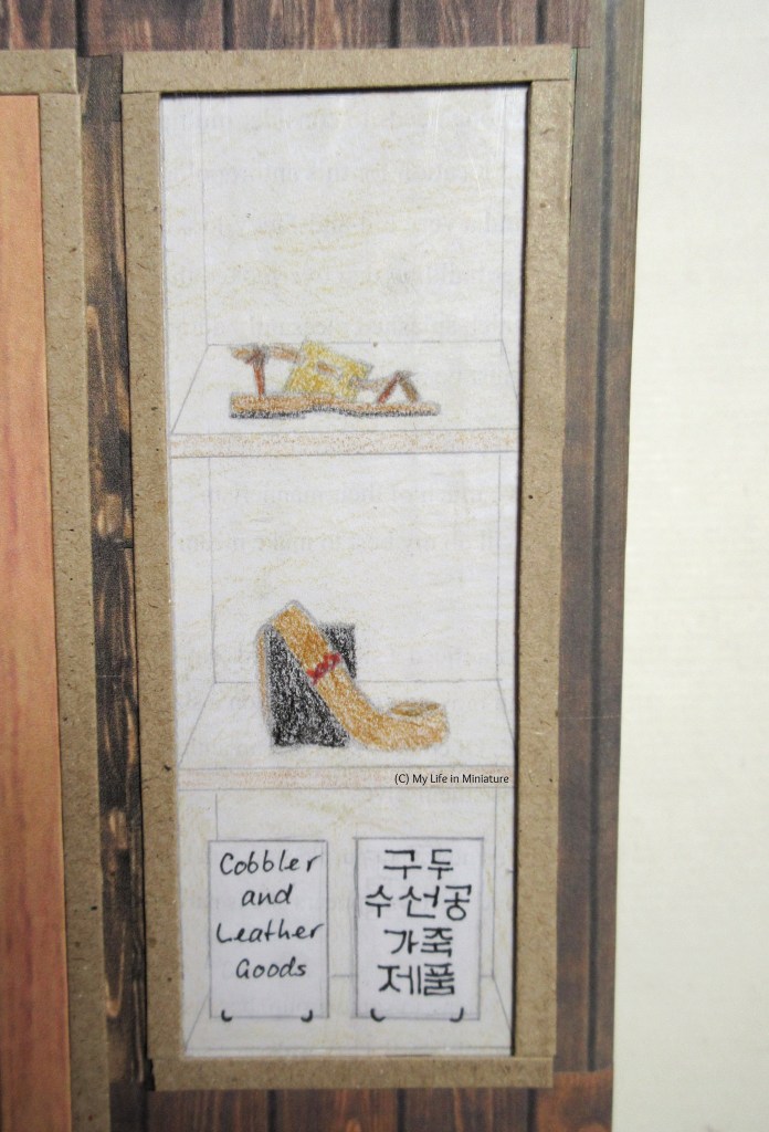 Close-up of the right-hand window. Similar to the left-hand window, the bottom shelf of the three has two signs that say 'Cobbler and Leather Goods', one in  English and one in Korean. 