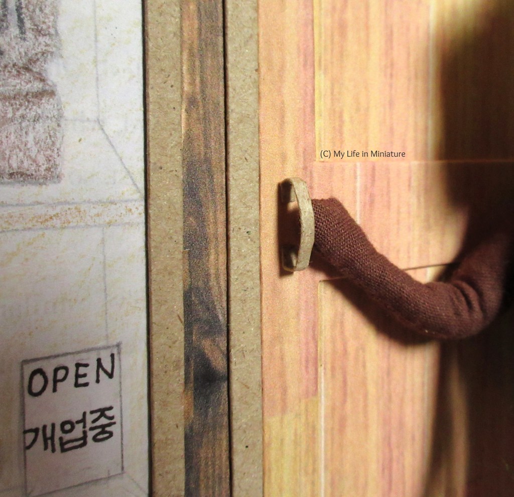 Close-up of the door handle. It is a vertical bar, made of the same slightly darker wood as the door and window frames. Hazel's hand is holding it so it's still visible to the camera. 