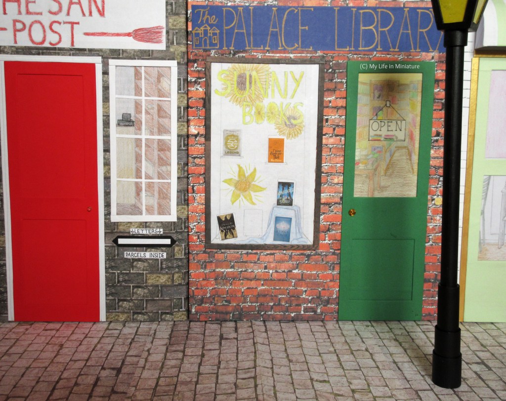 Another broad shot of the street, this time of the other side. Whesan Post is on the left, with The Palace Library in the middle and Fierro's Bakery on the right. Towards the foreground on the right is a black lamppost. 