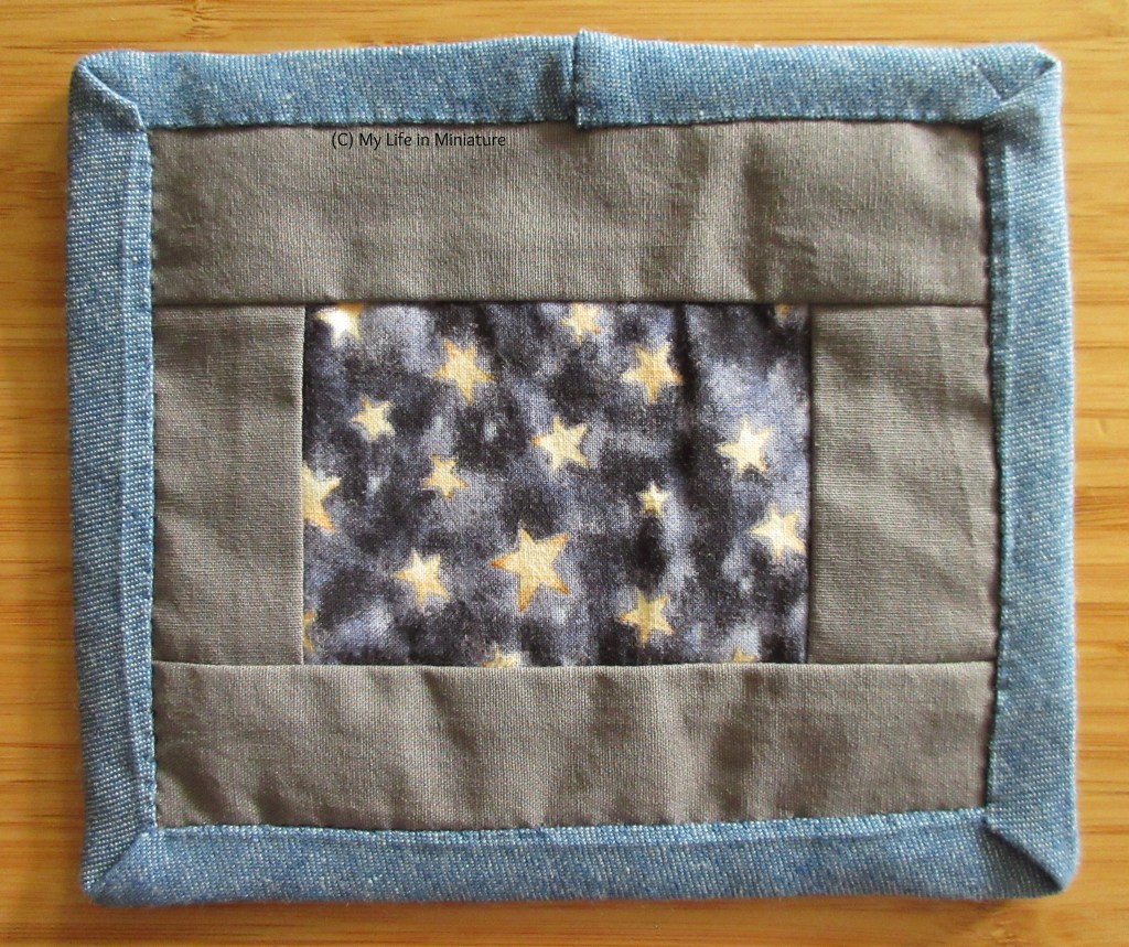 The very small rectangular quilt, still on a wood surface. The denim blue binding is stitched down to this side now. The patchwork on this side has a rectangle of starry fabric in the middle with plain grey rectangles around it.