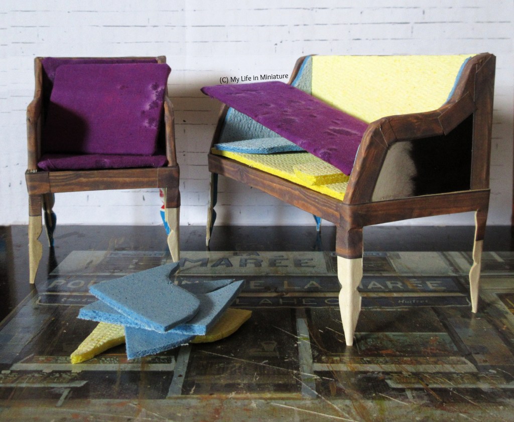 A matching couch and armchair, both half-complete, stand in front of a white brick background. They are both vaguely old-fashioned in style, though it's hard to tell because they are unfinished. There's dark wood and purple fabric happening, though.