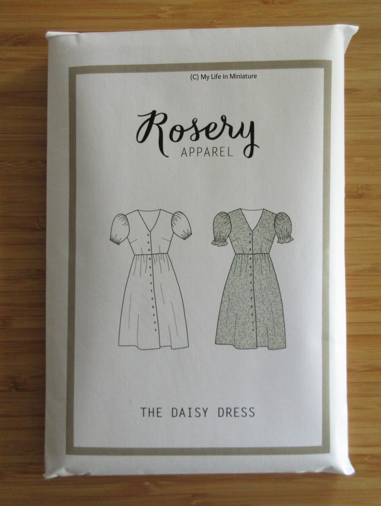 The pattern envelope of Rosery Apparel's Daisy dress pattern. It has an olive green border, the logo at the top, the dress diagram in the centre, and the text at the bottom. The dress is a button-down, V-necked midi-length, with puffed sleeves. 