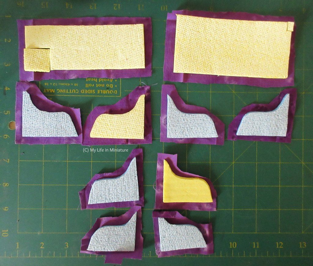 Rectangular and vaguely arm-of-a-chair-shaped sponges are laid out on purple fabric of corresponding shapes, on a green cutting mat. There are two rectangles, and the rest are vaguely arm-of-a-chair-shaped, in various sizes. 