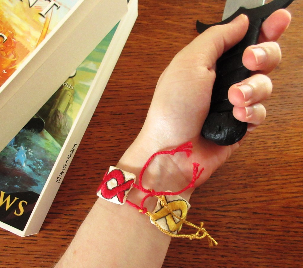 Another birds-eye shot over the wood surface. The tie end of the bracelet is visible, as the inside of the wrist is shown. The hand is holding a prop dagger, and the two books are to the top left of shot. 