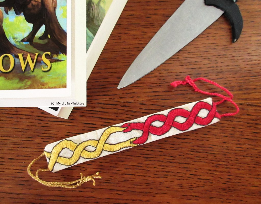 The bracelet is flat-laid on the wood surface, embroidered side up this time. The snake design is more visible - the yellow snake is intertwined with itself on the left side of the bracelet, while the red snake mirrors it on the right. They are both swallowing the other's tail in the middle of the bracelet. 