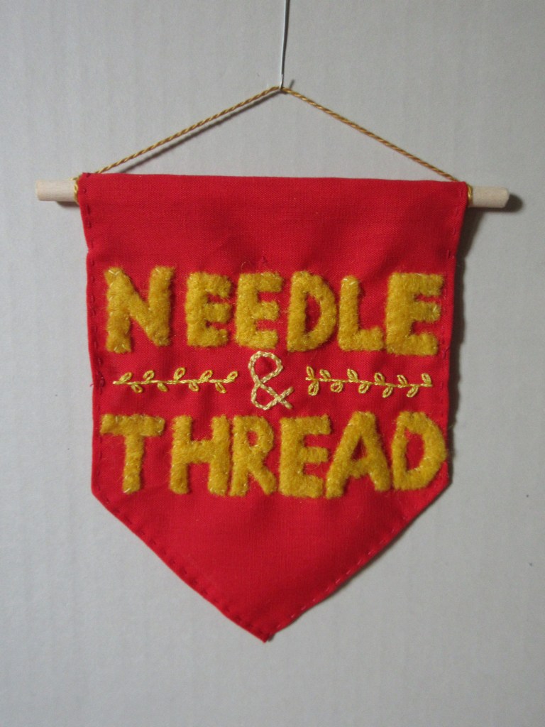 Close-up of the banner, hanging by a gold cord on a grey wall. The words 'Needle' and 'Thread' are appliquéd felt, and the ampersand is embroidered. It sits in-between the two words, and there are leafy branches embroidered on either side. 
