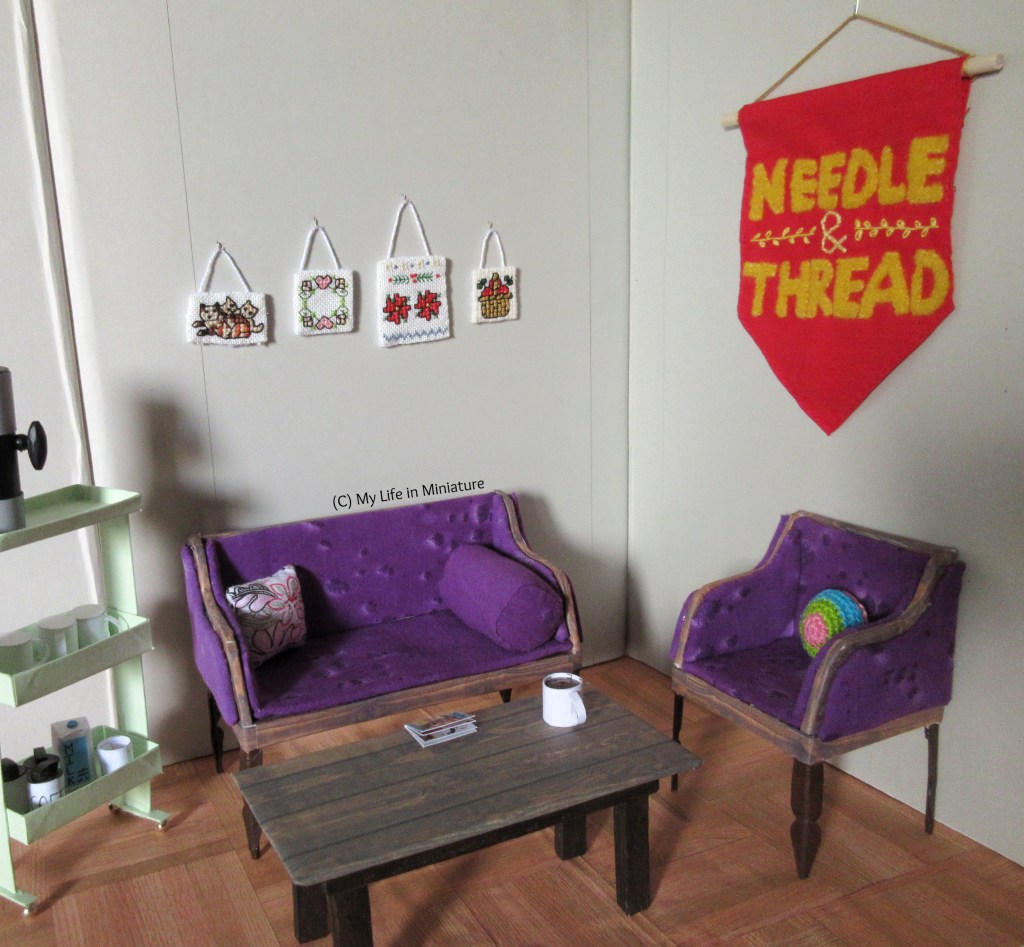 The Sit and Stitch corner in Needle & Thread in its entirety. Hanging on the wall to the left is a red and yellow banner that says 'Needle & Thread'. Below it is the purple armchair, with the purple couch against the adjoining wall. The coffee trolley is to the left of shot, and the wooden coffee table has a magazine and a mug on it.