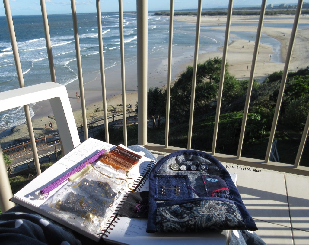 The author sits on a chair on a different balcony, overlooking a beach, the sea, and an island in the distance. On her lap is the same open sketchbook, with the fabrics, threads, and travel sewing kit open as before. 