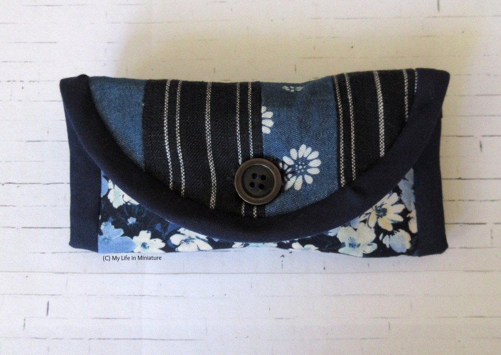 A small rectangular pouch sits on a white brick background. It buttons closed in the centre front. The flap is vertically striped in two different fabrics, and the fabric under the flap is different as well.