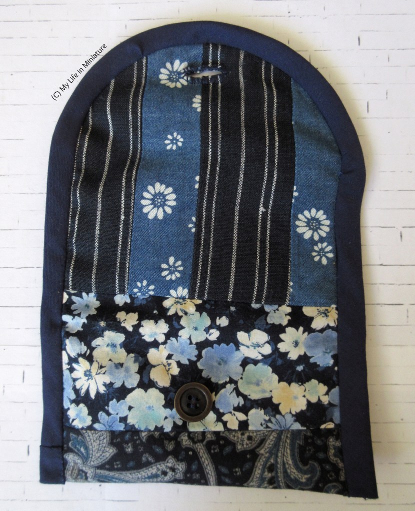 The travel sewing kit is open flat, outside up, on a white brick background. The top half is striped with a dark blue/white stripe fabric and a denim/white flower fabric. The bottom half is mostly a dark blue/blue and white floral fabric, with a little bit of dark blue paisley poking out. The outside edge is bound in dark blue bias binding.