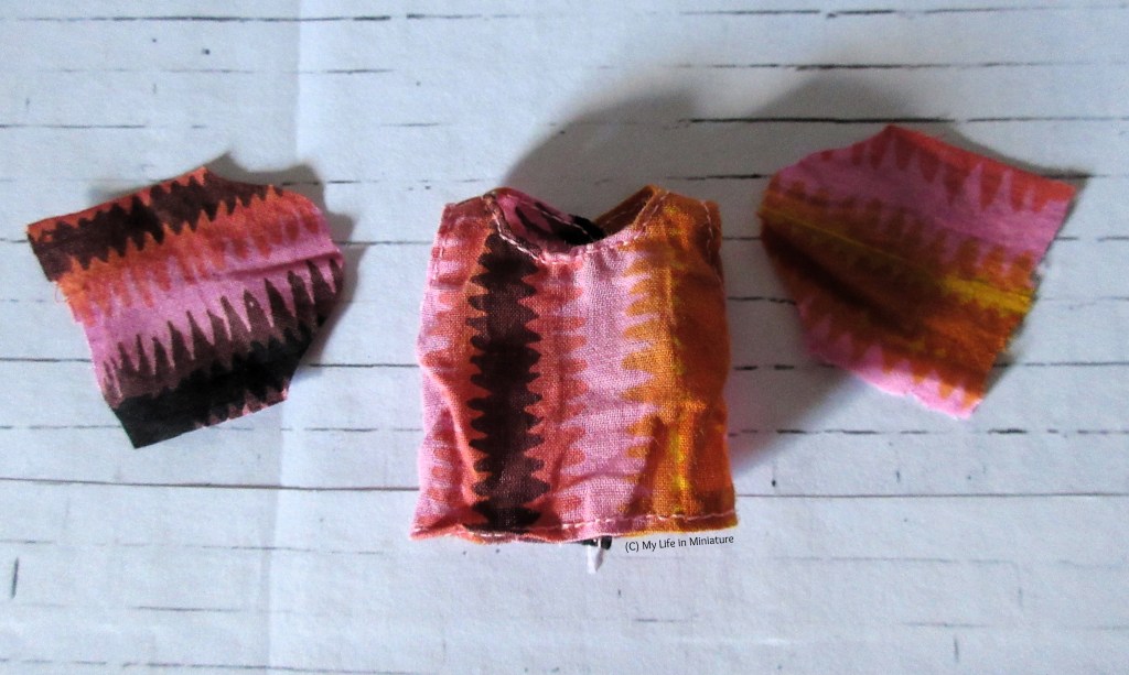 A pink, brown, and orange sleeveless shirt of Tiffany's is laid flat on a white brick surface. Next to its armholes are two flat sleeve shapes in the same fabric, ready to be assembled. 