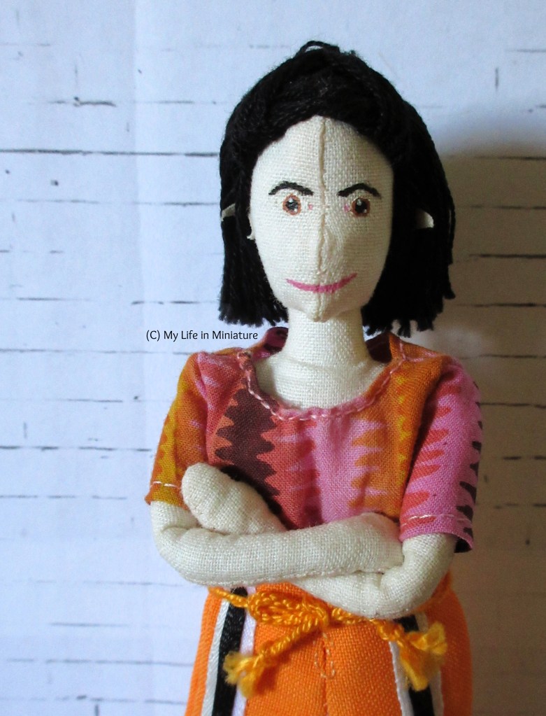 Tiffany stands in front of a white brick background, arms crossed over her chest, smiling at the camera. She is wearing the pink/orange/brown shirt from before, which now has short sleeves. (She's also wearing orange pants with black and white stripes.)