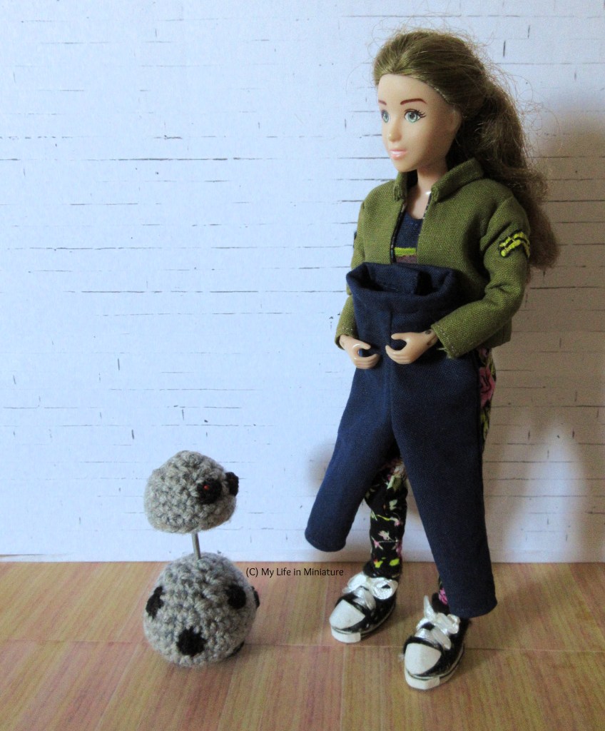 Sarah stands against a white brick background, holding a pair of navy pants up to herself. She's wearing black floral pants, and a navy striped shirt under a khaki jacket. Chip stands at Sarah's feet, their head elevated in interest at the pants. 