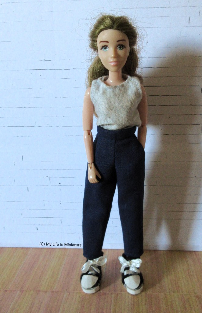 Sarah stands against a white brick background, facing the camera with a smile. She wears a sleeveless grey jersey top with the navy pants from before and black shoes. She has one hand in a pocket, and the other loose in front of her. 